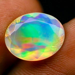 May include: An oval-shaped faceted opal gemstone displaying a vibrant play-of-color with iridescent flashes of green, blue, yellow, and orange across its translucent surface. The stone features a polished, multi-faceted cut that reflects light.
