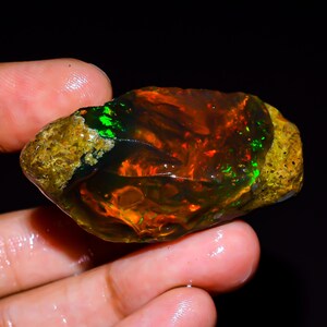 May include: A rough, uncut opal gemstone with a vibrant display of color. The opal features flashes of green, orange, and red within a dark, translucent body. The outer edges have a rough, golden-brown matrix.