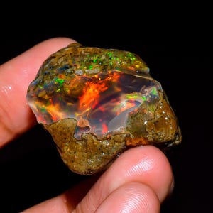 Op de afbeelding: Een ruwe, ongeslepen opaal edelsteen met levendige kleuren. De opaal toont een vurige mix van oranje, rood en groene tinten, met een doorschijnende kwaliteit. De steen heeft een ruwe, getextureerde buitenkant en wordt in een hand gehouden tegen een zwarte achtergrond.