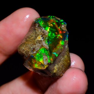 Op de afbeelding: Een ruwe opaal edelsteen met levendige kleuren. De opaal toont flitsen van groen, oranje en blauw tegen een bruine matrix. De edelsteen wordt in een hand gehouden, wat de natuurlijke, ruwe schoonheid benadrukt.