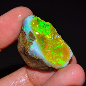 May include: A colourful, raw opal gemstone with a striking display of colour. The opal features a mix of bright green, yellow, and blue hues, with a rough, natural exterior. The stone is held in a hand, showcasing its unique, iridescent qualities.