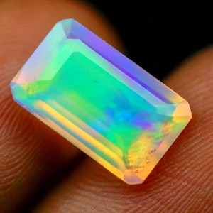 May include: A rectangular, emerald-cut opal gemstone displaying a vibrant play-of-color with iridescent flashes of green, blue, and yellow throughout its translucent, polished surface, showcasing a distinct faceted edge and a clear, glassy appearance.