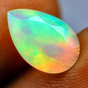 May include: A pear-shaped faceted opal gemstone displaying a vibrant play-of-color with iridescent flashes of neon green, yellow, and soft pink across its translucent surface. The gemstone features a polished, multi-faceted cut that reflects light.