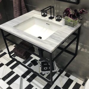 May include: A modern bathroom vanity with a white marble countertop and a black metal frame. The vanity has a white sink, a black faucet, and a lower shelf. Decorative flowers in a glass vase sit on the countertop. The floor has a black and white pattern.