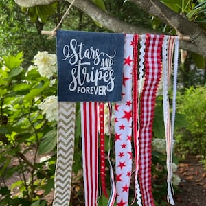 May include: A denim banner with the text "Stars and Stripes Forever" hanging from a branch. The banner is decorated with red, white, and blue ribbons.
