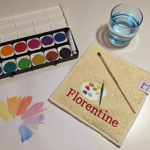 Könnte beinhalten: Ein Aquarellfarben-Set mit einer Vielzahl von Farben in einer Plastikpalette. Die Palette ist geöffnet und die Farben sind sichtbar. Es gibt einen kleinen Pinsel und ein Glas Wasser auf einem beigefarbenen Handtuch mit dem aufgestickten Wort "Florentine".