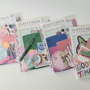 May include: Four clear plastic pouches filled with colorful ephemera, including stickers, paper cutouts, and a green pen. Each pouch has a "GUEST CHECK" header. The pouches contain various designs, including cherries, a smiley face, and the words "Sun City" and "Good Time."