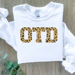 Puede incluir: Sudadera blanca con las letras "OTD" impresas en negro con un estampado de leopardo.