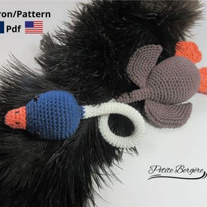 Peut inclure: Un hochet d'oiseau crocheté à la main avec une tête bleue, un bec orange et un cou blanc. Le hochet a un corps gris avec des accents orange et gris. Le fond présente des plumes noires. Le texte "Patron/Pattern Pdf" est visible.