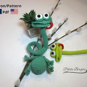 May include: Handmade green crocheted chameleon toy with white eyes and a red tongue. The reptile has a long neck and tail, with small arms and legs. The image also includes a small succulent plant in a glass container and the text "Patron/Pattern Pdf".