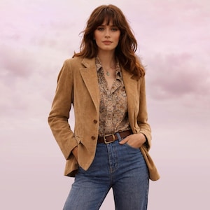 Puede incluir: Chaqueta de pana color canela con cierre de dos botones, combinada con una camisa estampada y vaqueros azules. Un cinturón de cuero marrón y un collar completan el conjunto. El fondo es un cielo rosa y morado.