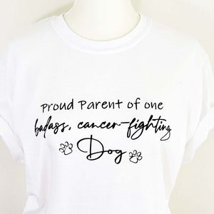 May include: White t-shirt with black text that reads "Proud Parent of one badass, cancer-fighting Dog" with paw prints on either side of the word "Dog".