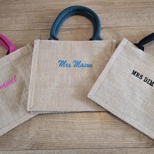 May include: Three square jute tote bags with sturdy handles. The left bag has pink handles and the name 'Sanel' in pink embroidery. The center bag has teal handles and 'Mrs Mason' in teal. The right bag has black handles and 'MRS DIMOND' in black.