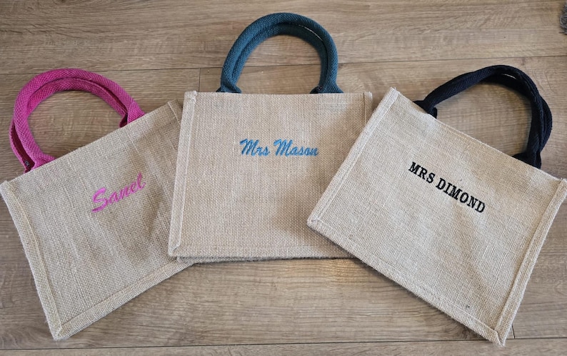 May include: Three square jute tote bags with sturdy handles. The left bag has pink handles and the name 'Sanel' in pink embroidery. The center bag has teal handles and 'Mrs Mason' in teal. The right bag has black handles and 'MRS DIMOND' in black.