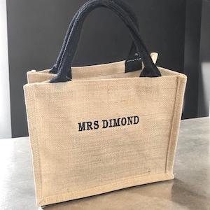 May include: A small rectangular tote bag made of tan woven jute fabric with two black fabric handles. The front of the bag features the text MRS DIMOND embroidered in black thread in a simple, bold, sans-serif font centered on the bag surface.