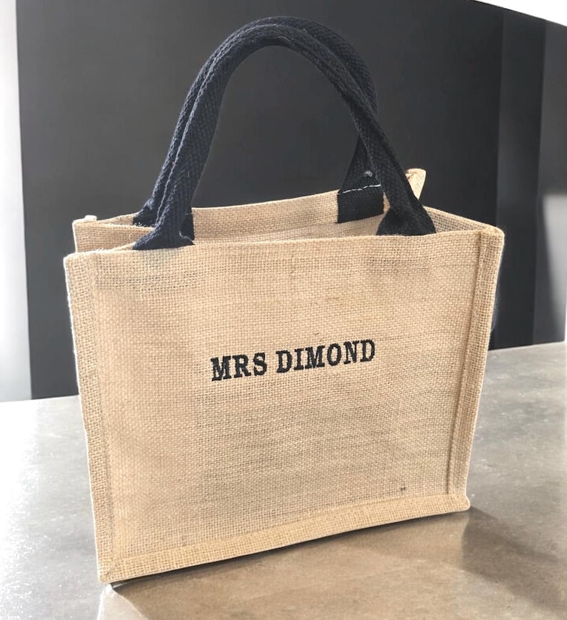 May include: A small rectangular tote bag made of tan woven jute fabric with two black fabric handles. The front of the bag features the text MRS DIMOND embroidered in black thread in a simple, bold, sans-serif font centered on the bag surface.