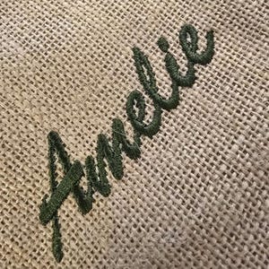 May include: A coarse, tan-colored burlap fabric featuring the name Amelie embroidered in a dark forest green thread. The embroidery is executed in a cursive script font, with the letters stitched directly onto the textured, woven material.