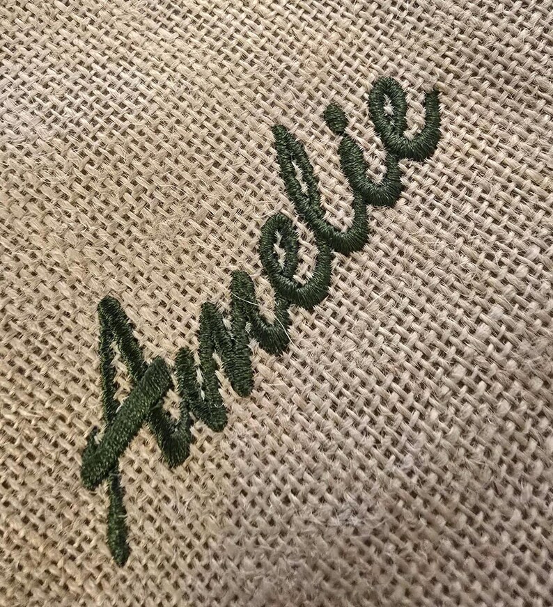 May include: A coarse, tan-colored burlap fabric featuring the name Amelie embroidered in a dark forest green thread. The embroidery is executed in a cursive script font, with the letters stitched directly onto the textured, woven material.