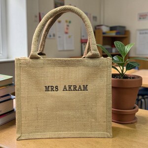 May include: A square-shaped tan burlap tote bag with two matching woven handles. The front of the bag features the text MRS AKRAM embroidered in dark brown thread. The bag has a structured rectangular base and a coarse, natural fiber texture.
