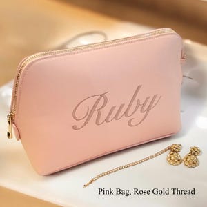 May include: A light pink cosmetic bag with a gold zipper and the name "Ruby" engraved on the front. A rose gold chain with two flower-shaped earrings is next to the bag. The text "Pink Bag, Rose Gold Thread" is at the bottom.