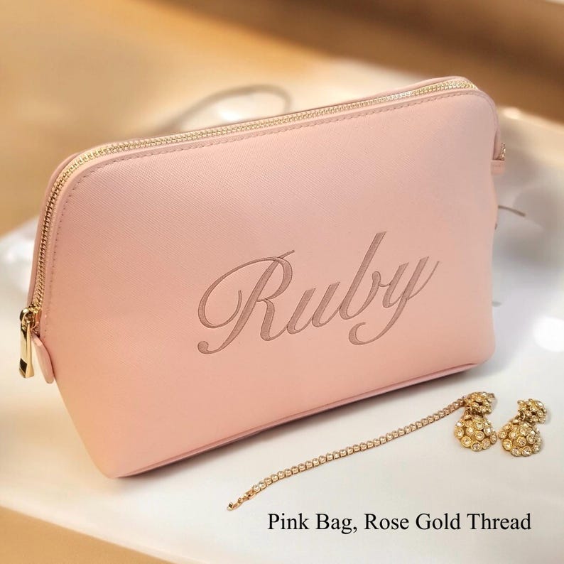 May include: A light pink cosmetic bag with a gold zipper and the name "Ruby" engraved on the front. A rose gold chain with two flower-shaped earrings is next to the bag. The text "Pink Bag, Rose Gold Thread" is at the bottom.