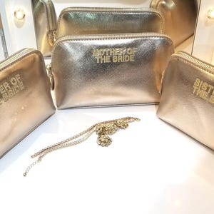 May include: Gold-colored cosmetic bags with the text "Sister of the Bride" and "Mother of the Bride" in gold lettering. A gold-colored necklace with a floral design is also present. The bags have a zipper closure.