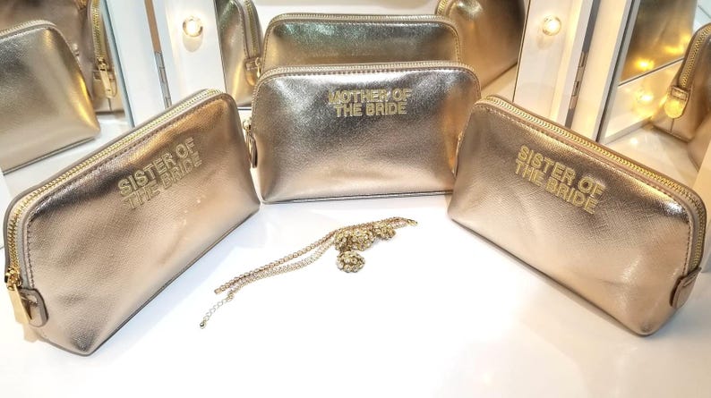 May include: Gold-colored cosmetic bags with the text "Sister of the Bride" and "Mother of the Bride" in gold lettering. A gold-colored necklace with a floral design is also present. The bags have a zipper closure.