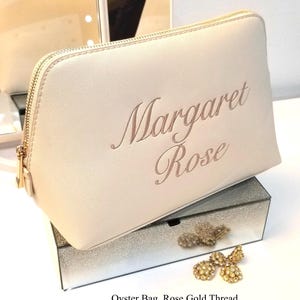 May include: A light beige oyster bag with a gold zipper and the name "Margaret Rose" embroidered in rose gold thread. The bag is displayed on a mirrored box with gold-colored jewelry.