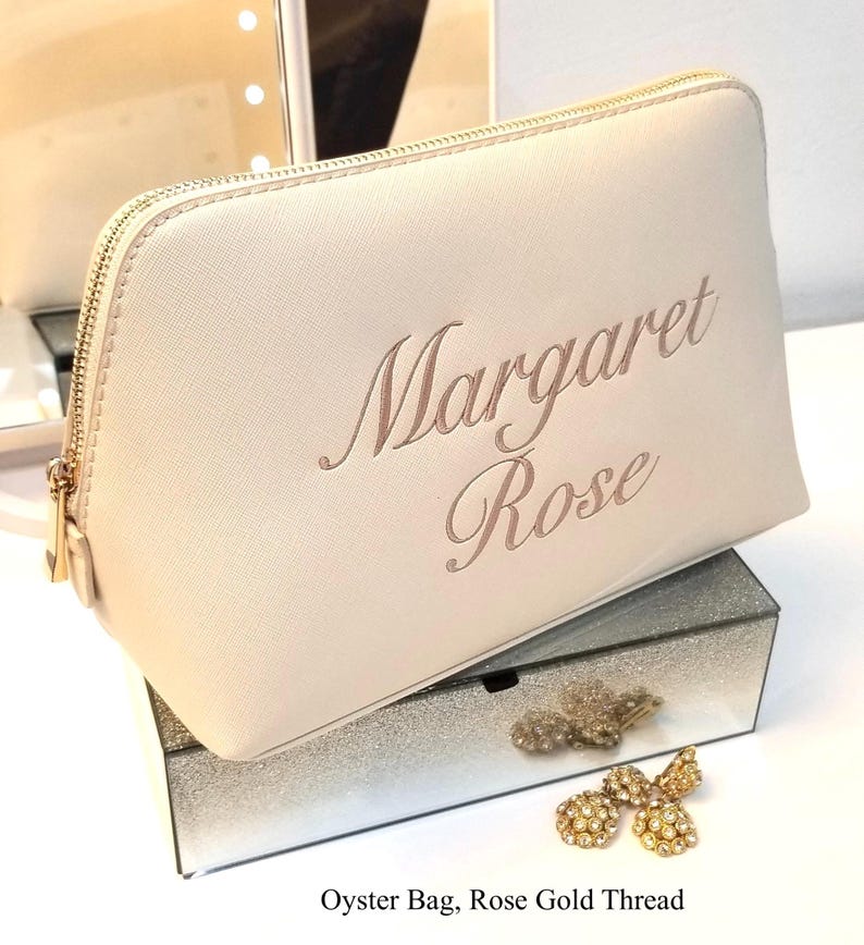 May include: A light beige oyster bag with a gold zipper and the name "Margaret Rose" embroidered in rose gold thread. The bag is displayed on a mirrored box with gold-colored jewelry.