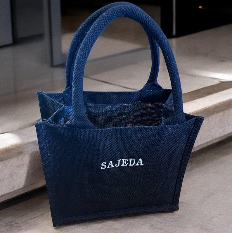 May include: A small, dark blue tote bag made of a textured woven fabric. The bag features two matching top handles and the name SAJEDA embroidered in white capital letters on the front center of the exterior panel of the open-top bag.