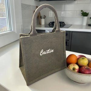 May include: A small, olive green jute tote bag with two sturdy top handles. The front of the bag features the name Caitlin embroidered in white cursive script. The bag has a structured, rectangular shape with a textured, woven fabric finish.