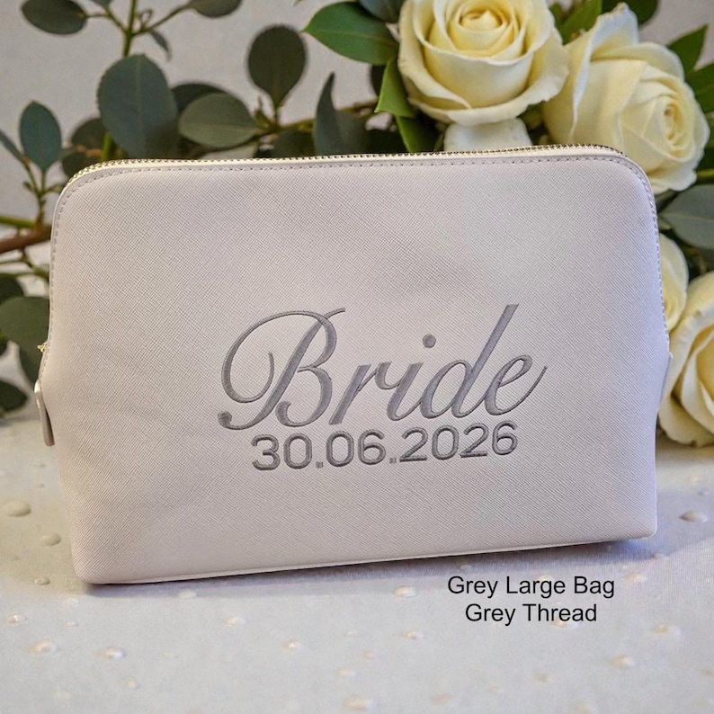 Może przedstawiać: Jasnoszara kosmetyczka z wyhaftowanym słowem "Bride" szarą czcionką, wraz z datą "30.06.2026". Torba ma zapięcie na zamek błyskawiczny i jest prezentowana na tle białych r&oacute;ż i zieleni.