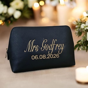 May include: A black cosmetic bag with a gold zipper and gold embroidered text that reads "Mrs Godfrey" and the date "06.08.2026". The bag is sitting on a light-colored surface.