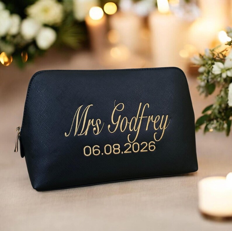 May include: A black cosmetic bag with a gold zipper and gold embroidered text that reads "Mrs Godfrey" and the date "06.08.2026". The bag is sitting on a light-colored surface.