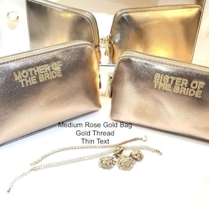 May include: Rose gold cosmetic bags with gold thread text that reads "MOTHER OF THE BRIDE" and "SISTER OF THE BRIDE". The bags have a zipper closure and are displayed with gold jewelry.