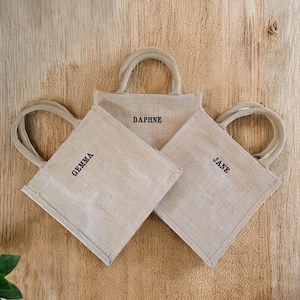 May include: Three square beige jute tote bags with rounded handles. Each bag features a name embroidered in black thread on the front: Gemma, Daphne, and Jane. The bags are arranged in a cluster, showcasing their natural woven texture.