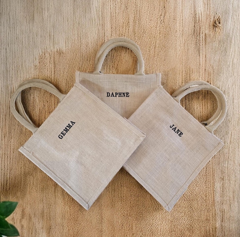 May include: Three square beige jute tote bags with rounded handles. Each bag features a name embroidered in black thread on the front: Gemma, Daphne, and Jane. The bags are arranged in a cluster, showcasing their natural woven texture.