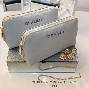 May include: Two medium gray cosmetic bags with gold zippers, one labeled "GLAMMY" and the other "CHELSEY". The bags sit on a mirrored box with jewelry, including gold earrings and a rhinestone bracelet. The text "MEDIUM GREY BAG WITH GREY TEXT" is at the bottom.