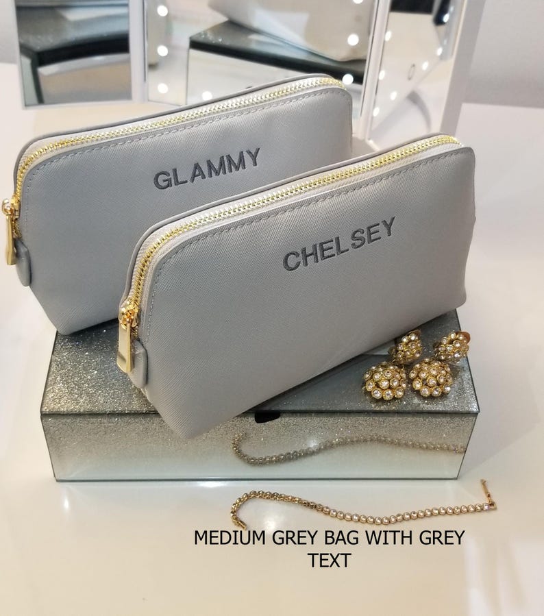 May include: Two medium gray cosmetic bags with gold zippers, one labeled "GLAMMY" and the other "CHELSEY". The bags sit on a mirrored box with jewelry, including gold earrings and a rhinestone bracelet. The text "MEDIUM GREY BAG WITH GREY TEXT" is at the bottom.