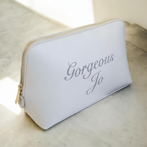 May include: A light gray cosmetic bag with a gold zipper and the text "Gorgeous Jo" in cursive. The bag is rectangular with rounded corners and sits on a light-colored surface.
