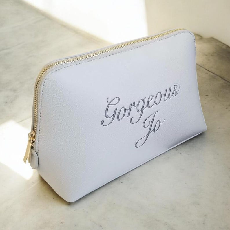 May include: A light gray cosmetic bag with a gold zipper and the text "Gorgeous Jo" in cursive. The bag is rectangular with rounded corners and sits on a light-colored surface.