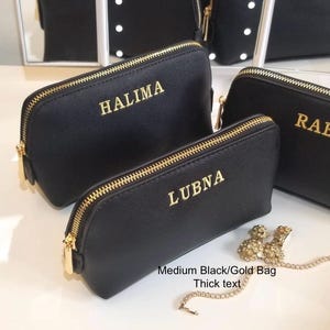 May include: Black cosmetic bags with gold zippers and personalized gold lettering. The bags are in various sizes, with the names "HALIMA", "LUBN", and "RAB" displayed. A small gold-colored jewelry piece is also visible.