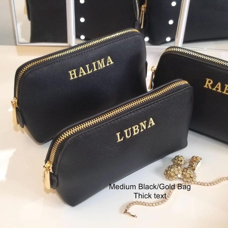 May include: Black cosmetic bags with gold zippers and personalized gold lettering. The bags are in various sizes, with the names "HALIMA", "LUBN", and "RAB" displayed. A small gold-colored jewelry piece is also visible.