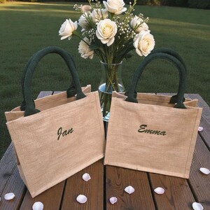 May include: Two rectangular tan jute tote bags with dark green handles. Each bag features a name embroidered in dark green cursive script on the front, with one reading Jan and the other reading Emma. The bags are positioned on a wooden surface.