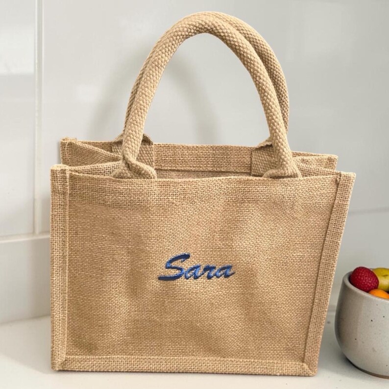 May include: A rectangular beige jute tote bag with two top handles. The front of the bag features the name Sara embroidered in a dark blue script font. The bag has a structured design with visible woven texture throughout the natural fiber material.