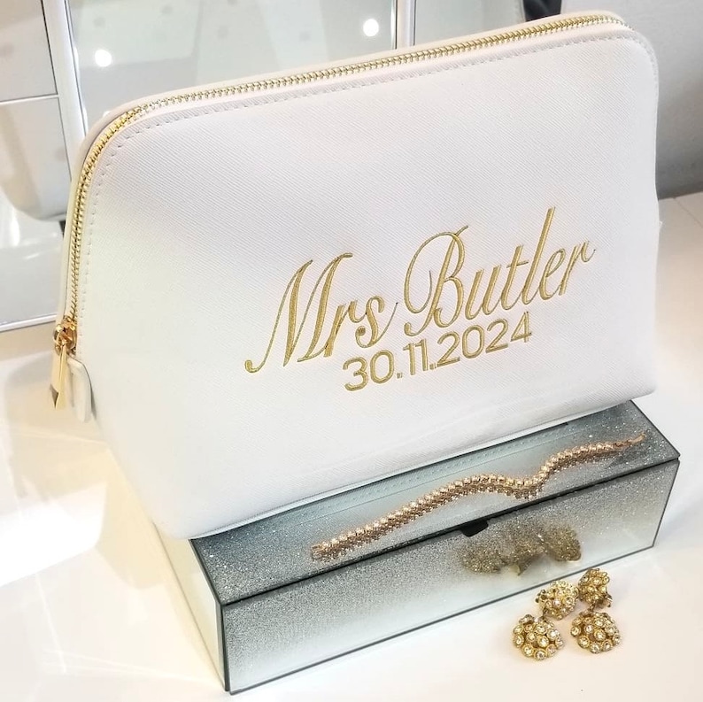 Może przedstawiać: Biała kosmetyczka ze złotym zamkiem błyskawicznym i haftowanym tekstem "Mrs Butler 30.11.2024". Torba spoczywa na lustrzanym pudełku ze złotą bransoletką i kolczykami.