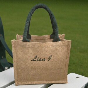 May include: A small square tote bag made of tan woven jute fabric. The bag features two dark green padded handles and the name Lisa J embroidered in dark green script lettering on the front center of the bag. The design is simple and structured.