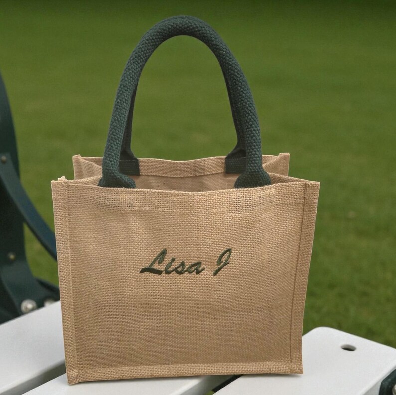 May include: A small square tote bag made of tan woven jute fabric. The bag features two dark green padded handles and the name Lisa J embroidered in dark green script lettering on the front center of the bag. The design is simple and structured.
