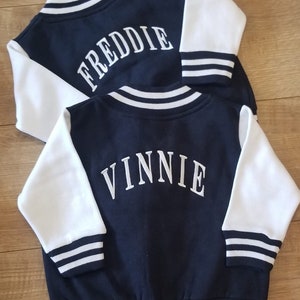May include: Two navy blue and white baseball jackets with white embroidered names on the back. The top jacket says "FREDDIE" and the bottom jacket says "VINNIE".