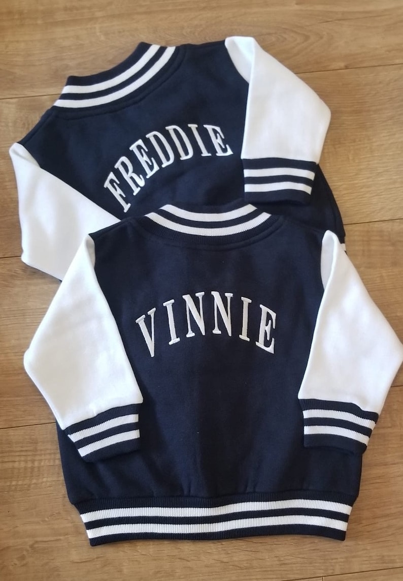 May include: Two navy blue and white baseball jackets with white embroidered names on the back. The top jacket says "FREDDIE" and the bottom jacket says "VINNIE".